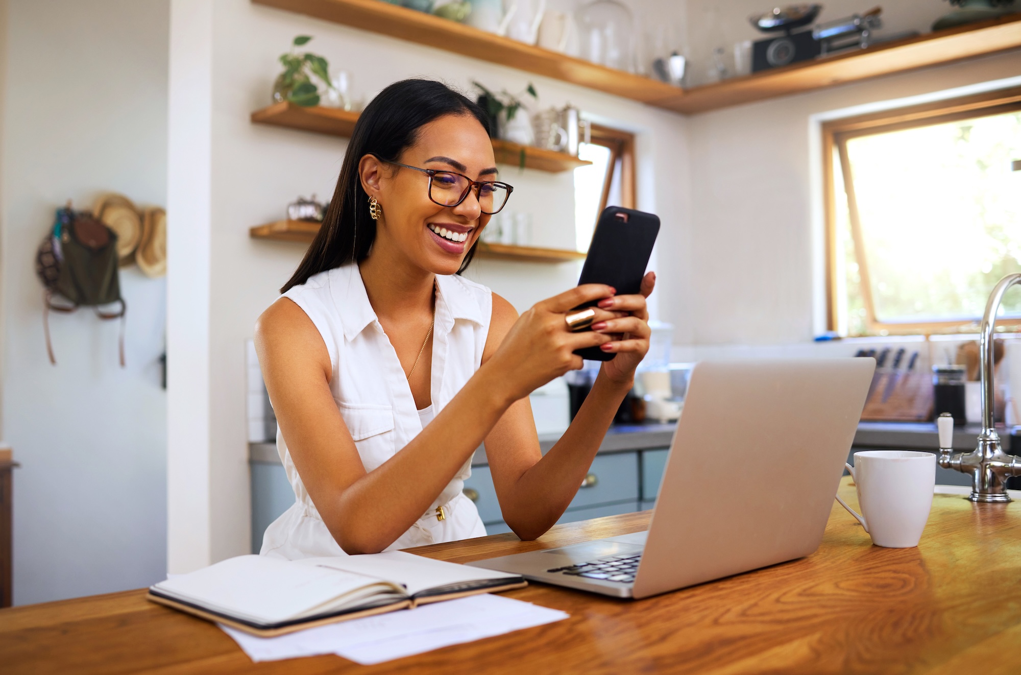 A woman sitting in front of a computer and holding up her phone and smiling at it