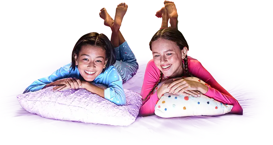 Two girls in pajamas, smiling and resting their arms on their pillows with their feet in the air.